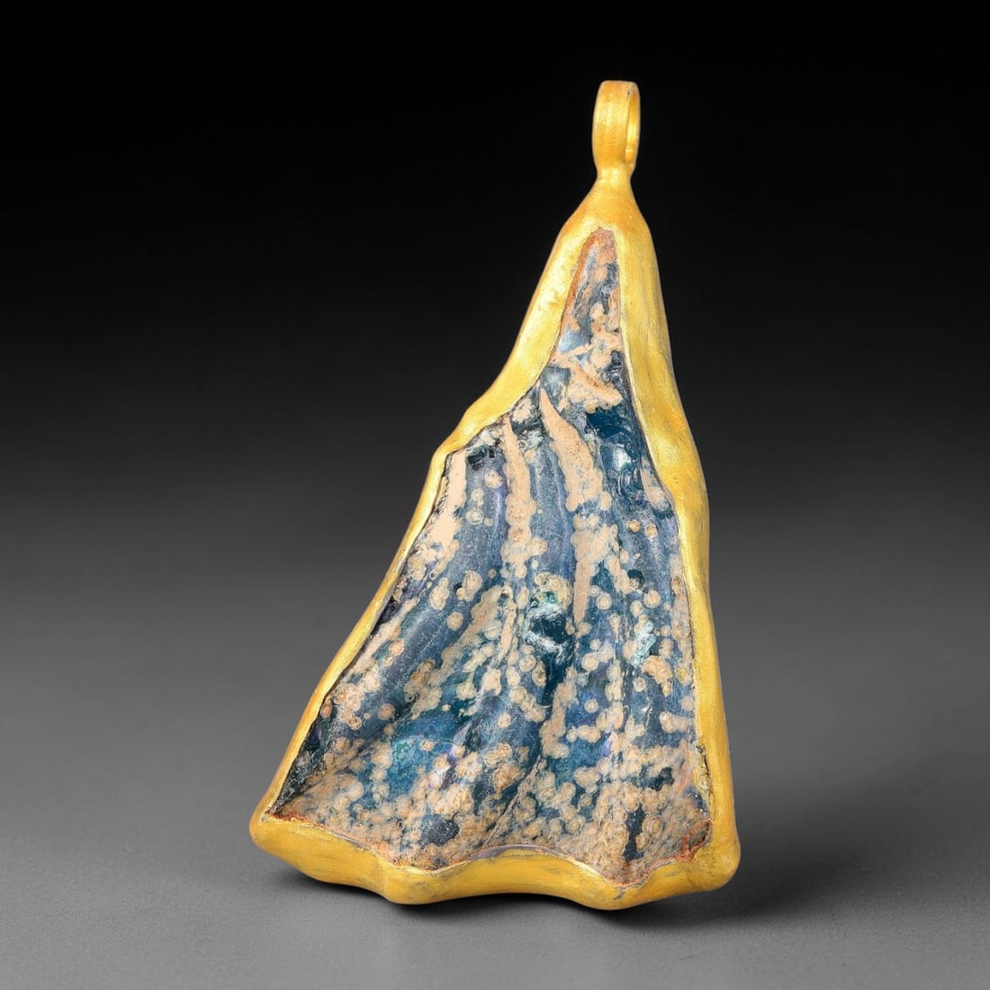 Ancient Roman Glass Slag in Modern Gilt-Edge Pendant, 50FY: This contemporary pendant is crafted from ancient Roman glass slag, featuring a fragmented, iridescent glass surface with weathered patina. The irregular form is bordered by a subtle gilt frame, added