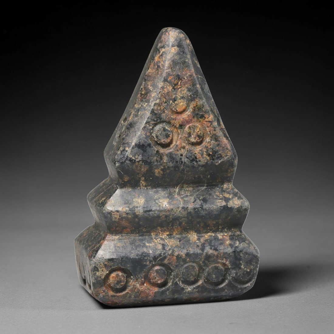 Ancient Medieval Mamluk Period Dark Stone Tiered Talisman (c.13th-16th Century CE), 61EC: This tiered pyramidal talisman is carved from dark mottled stone, decorated with drilled circular symbolic motifs across its surfaces. Aged earthy patina and soft surface wear develop over generations