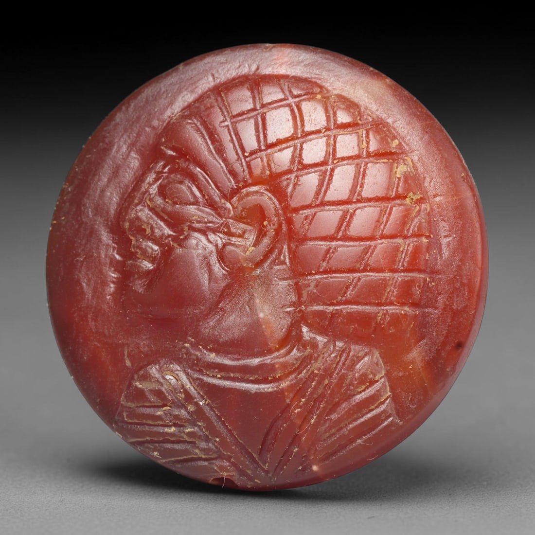 Ancient Medieval Early Islamic Period Carnelian Cameo Bead (c.7th-11th Century CE), 74EC: This circular pierced bead is crafted from natural carnelian, featuring a finely rendered cameo relief profile of an anthropomorphic bust with elaborate styled hair, a common motif within medieval Nea