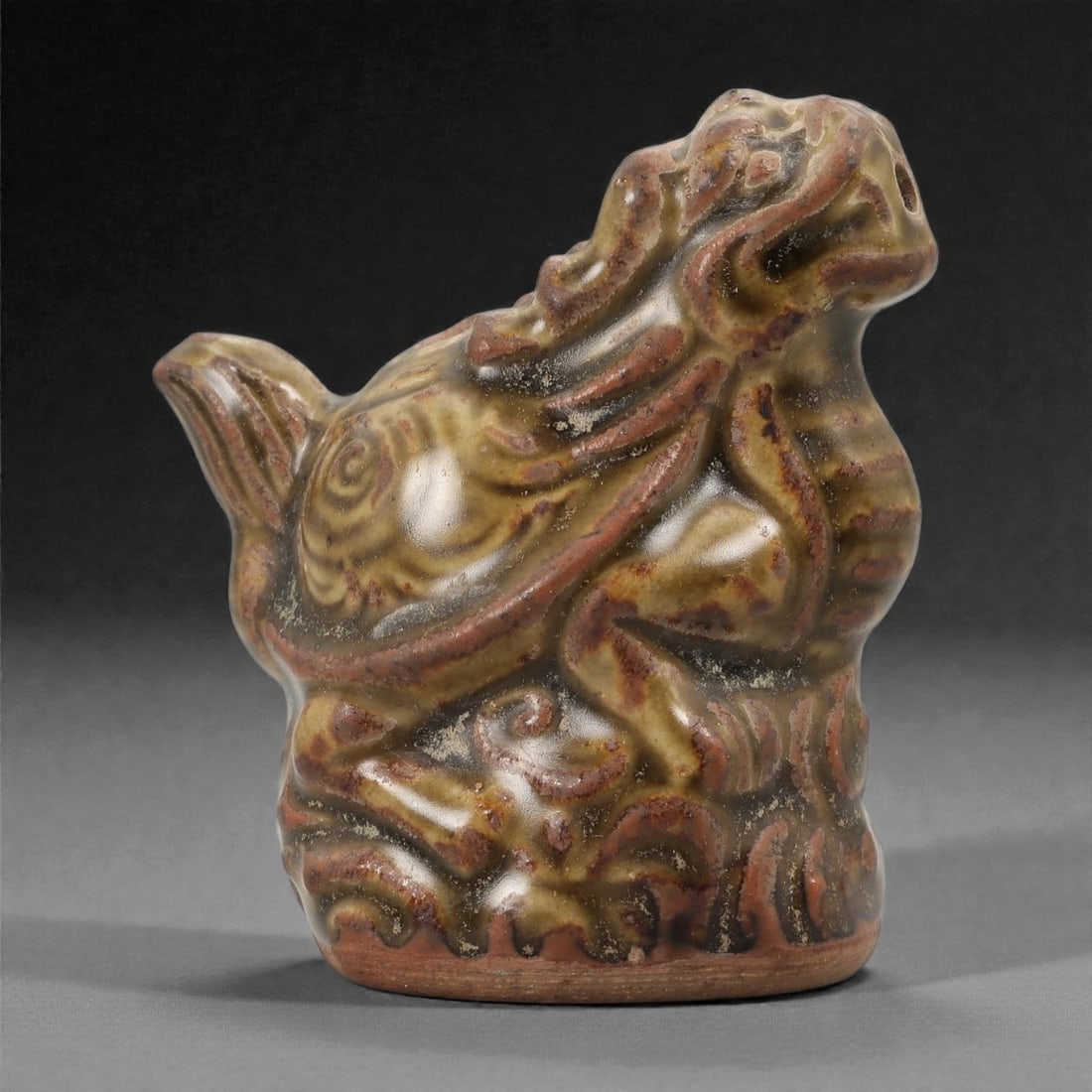 A Chinese Glazed Ceramic Bixi Figure, 78FG: Modeled from ceramic and covered with a mottled glaze in olive green, brown, and reddish tones, the figure represents bixi, the mythical creature combining the body of a tortoise with the head of a dr