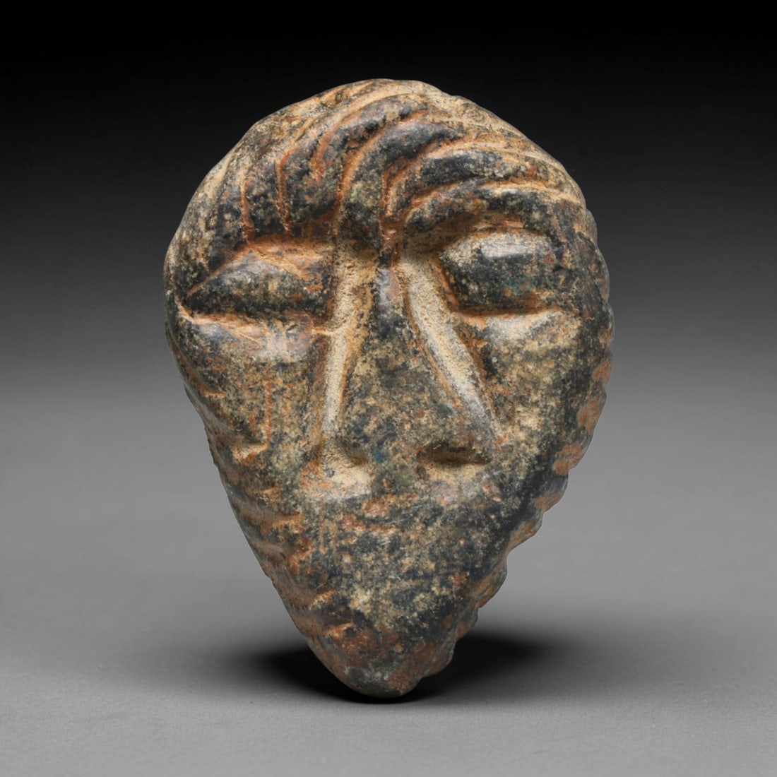 Ancient Mesopotamian Stone Maskette Amulet (c.2300-539 BCE), 43ED: This carved dark stone maskette amulet originates from Ancient Mesopotamia. Deep incised lines define facial features, stylised hair and facial hair following established regional iconographic convent