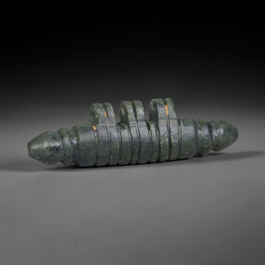 Ancient Near Eastern Dark Green Stone Ritual Talisman, 94ED: This antique dark green stone ritual talisman originates from the Ancient Near East. The elongated cylindrical artifact features segmented carved detailing, dual tapered terminals and three integrated