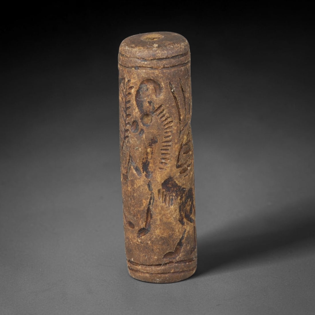 Ancient Mesopotamian Stone Cylinder Seal (c.2300-539 BCE), 67EF: This carved pale weathered stone cylinder seal originates from Ancient Mesopotamia. The cylindrical body features continuous incised imagery encircling the surface, framed by simple horizontal border 