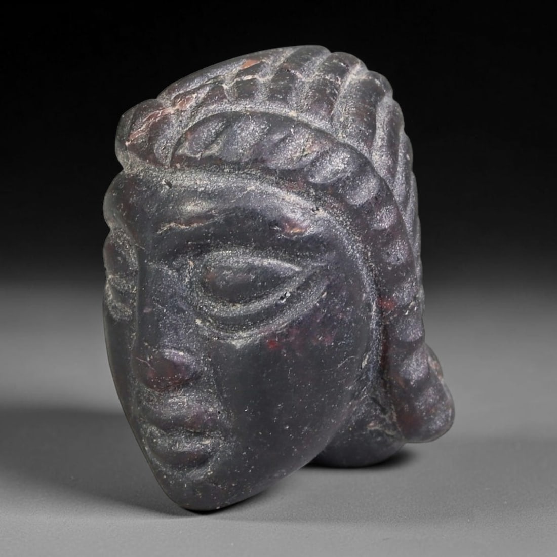 Antique Carved Dark Stone Human Face Sculpted Bead (Unpierced), 92FL: This antique carving is fashioned from dark mineral stone, rendered as a stylised human face bead without a suspension perforation. Intricate detailing defines facial features and textured coiffure, s