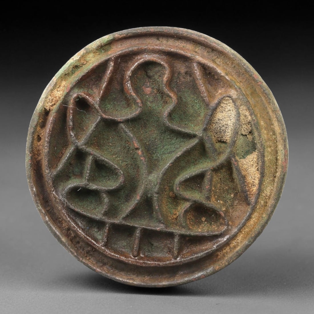 An Ancient Bactrian Bronze Stamp Seal, 54AG: A circular bronze stamp seal of the Bactrian period, with a raised relief design depicting a stylized central motif flanked by curvilinear elements, framed within a double border. The surface displays