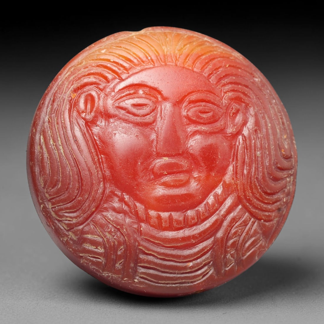 Ancient Medieval Early Islamic Period Carnelian Cameo Bead (c.7th-11th Century CE), 3ED: This circular pierced bead is crafted from natural carnelian, showcasing a raised cameo carving of a frontal anthropomorphic bust adorned with elaborate layered hair and decorated robes, typical of me