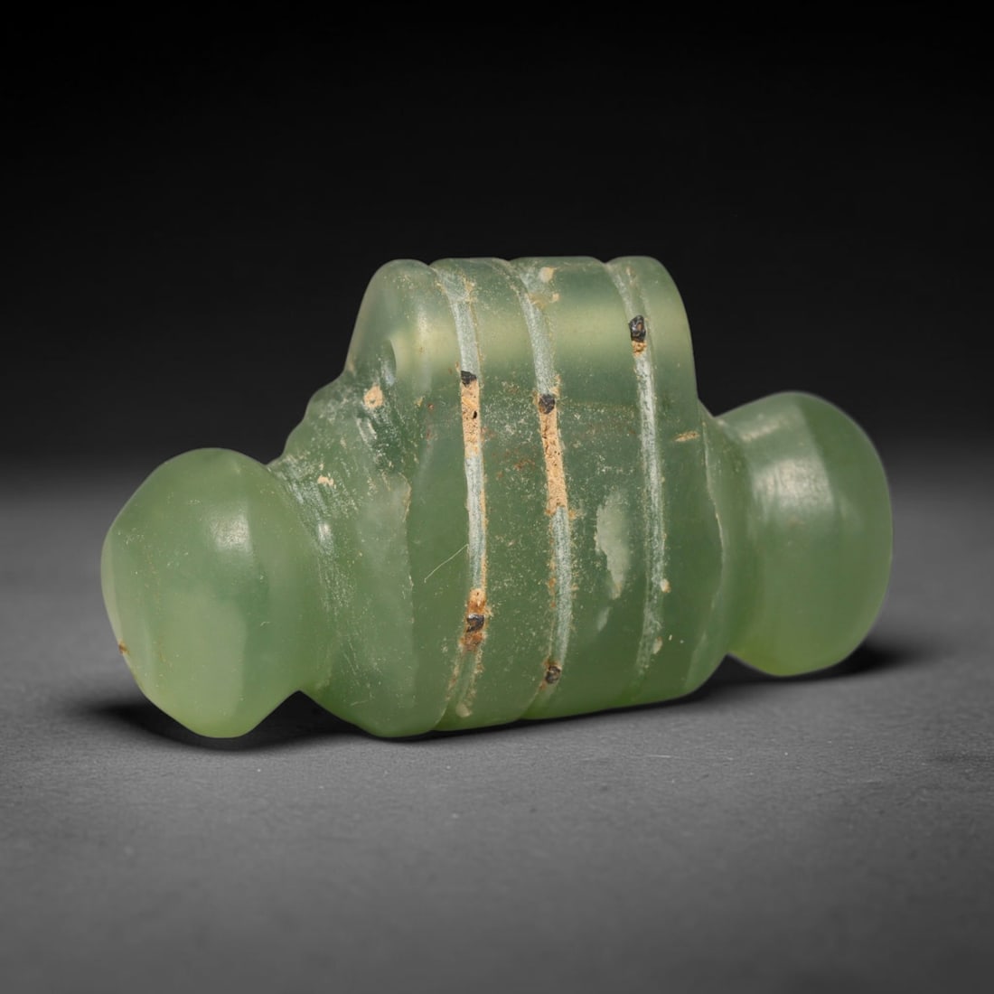 Ancient Sasanian Persian Carved Green Stone Amulet (c.224-651 CE), 27ED: This translucent pale green stone amulet originates from Ancient Sasanian Persian culture. The symmetrical stylised form features evenly spaced incised grooves encircling its central body, flanked by 