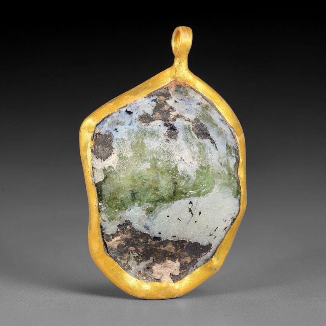 Ancient Roman Glass Slag in Modern Gilt-Edge Pendant, 60FY: This contemporary pendant is crafted from ancient Roman glass slag, featuring a fragmented, iridescent glass surface with weathered patina. The irregular form is bordered by a subtle gilt frame, added