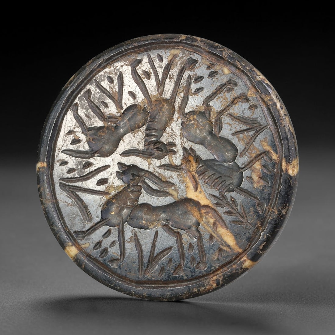 Ancient Syro-Hittite Dark Stone Intaglio Plaque (1200-800 BCE), 15EF: This circular stone intaglio plaque is carved from mottled dark stone, originating from Iron Age Syro-Hittite civilizations of Northern Syria and Eastern Anatolia. The surface is engraved with a compo