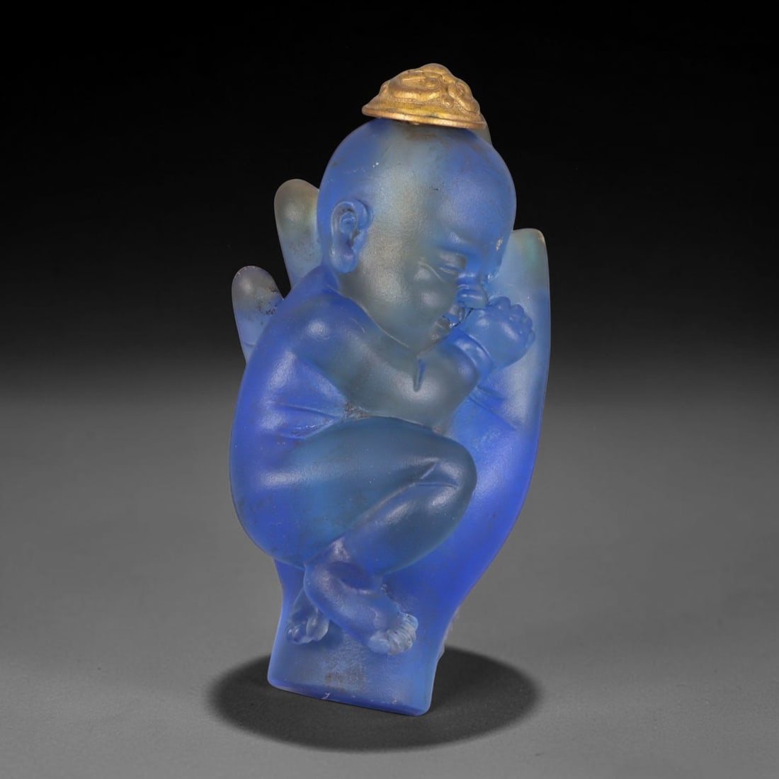 Vintage Chinese Liuli Glass Snuff Bottle - Baby in Deity Hand, 36AS: This snuff bottle is moulded from frosted translucent blue liuli glass, carved as a sleeping baby curled inside a Buddha hand, symbolizing divine protection. The piece features smooth rounded carving 