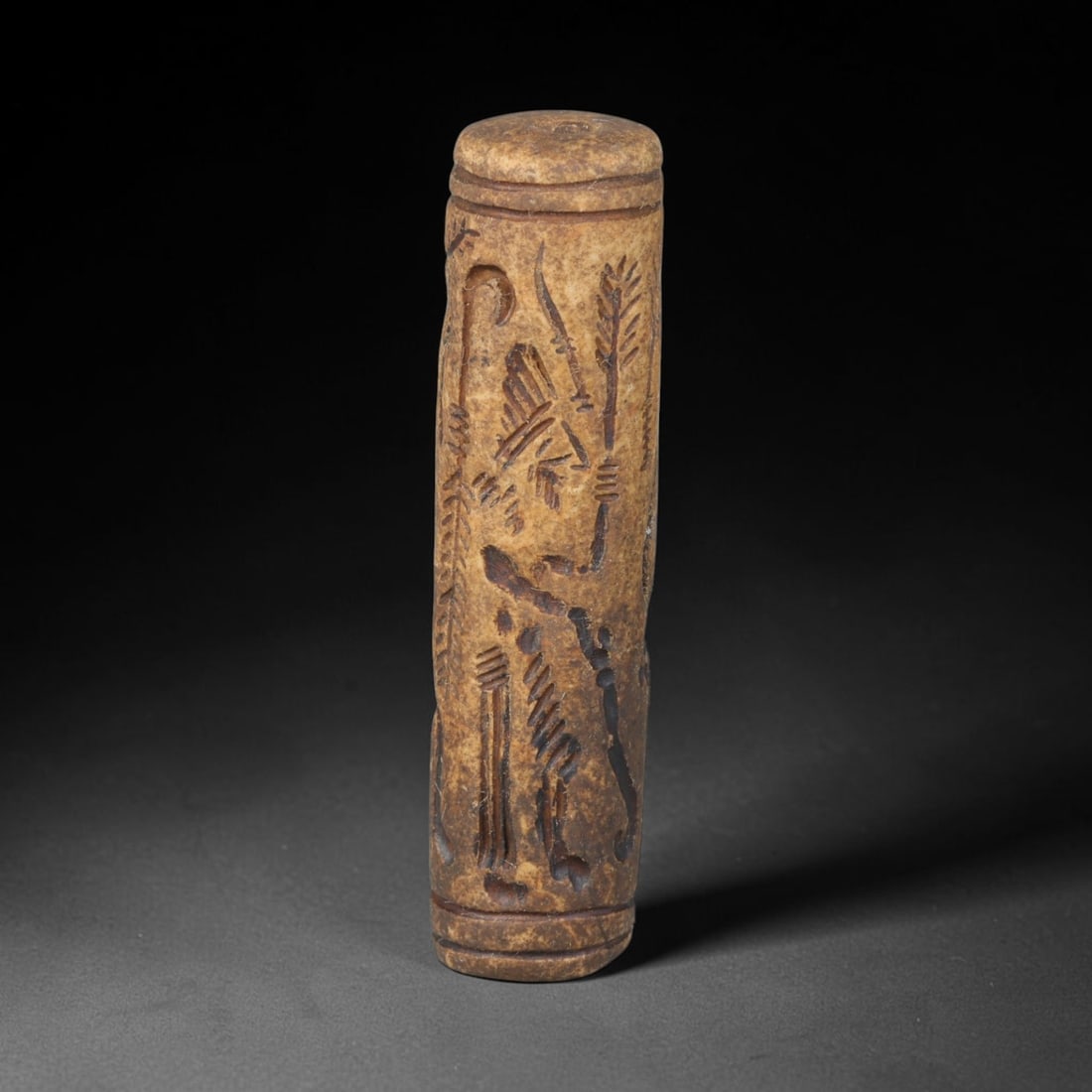 Ancient Mesopotamian Stone Cylinder Seal (c.2300-539 BCE), 63EF: This carved pale weathered stone cylinder seal originates from Ancient Mesopotamia. The cylindrical body features continuous incised imagery encircling the surface, framed by simple horizontal border 
