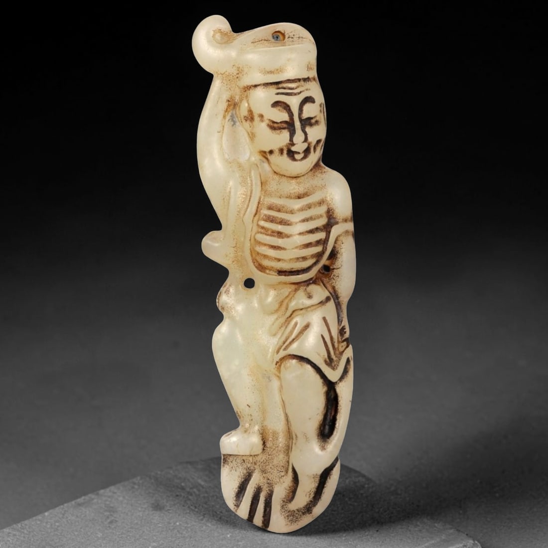 A Chinese Ancient Style Jade Stone Figural Pendant, 53FA: Carved from pale cream-colored stone with darker brown surface staining accentuating the incised details, the pendant depicts a standing human figure rendered in a simplified, elongated form. The figu