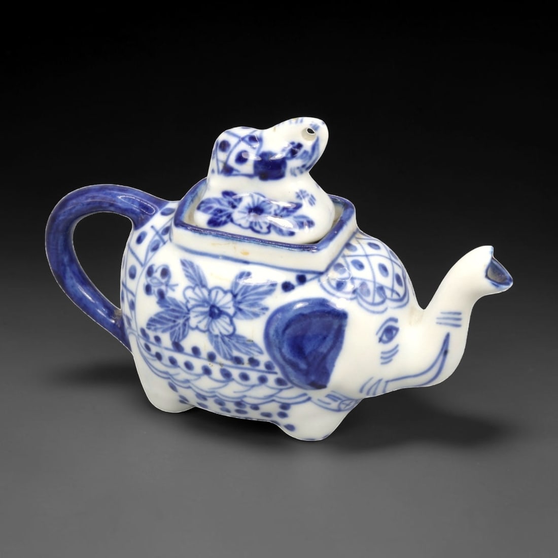 A Chinese Porcelain Elephant-Form Teapot, 20th Century, 46FK: Modeled in the form of a recumbent elephant, the teapot is constructed from white porcelain and decorated in underglaze cobalt blue. The elephant's raised trunk functions as the spout, while a looped 