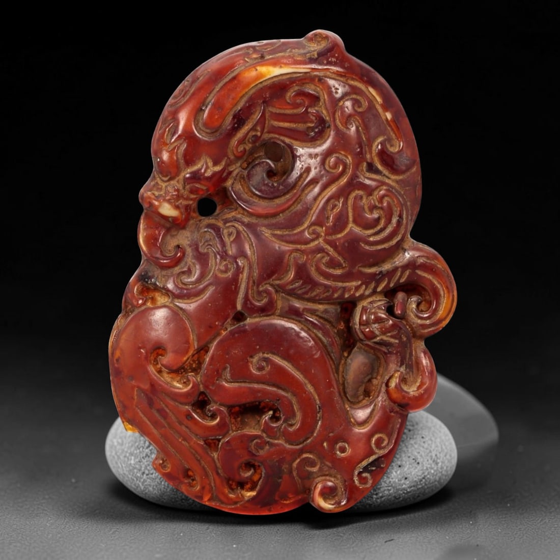 A Chinese Amber-Resin Openwork Plaque Pendant, 90FC: Molded and carved from translucent reddish-brown amber-colored resin with areas of lighter translucency and surface variation, the object is worked as an irregular plaque featuring a stylized mask-lik