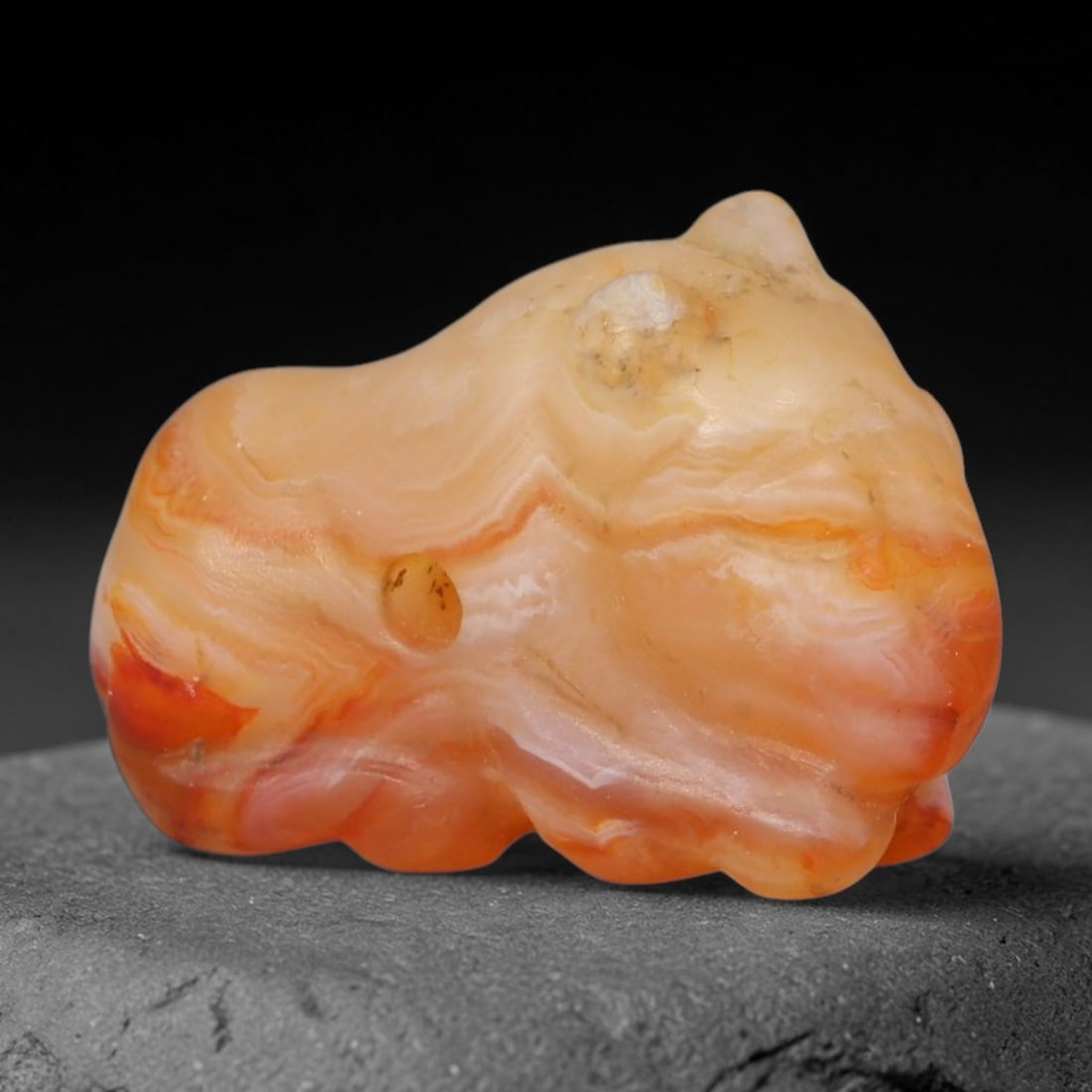 Ancient Western Asiatic Banded Agate Recumbent Feline Zoomorphic Amulet (1000-600 BCE), 28AB: Hand-carved translucent banded agate pendant formed into a stylized reclining cat/feline figure, featuring natural flowing orange, cream and pale red layered banding throughout the stone matrix. A nea