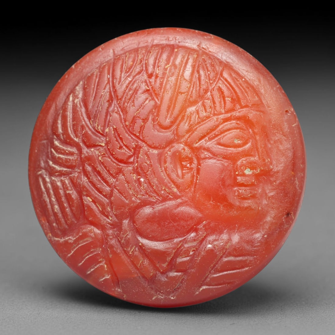 Ancient Medieval Early Islamic Period Carnelian Cameo Bead (c.7th-11th Century CE), 75EC: This circular pierced bead is crafted from natural carnelian, featuring a finely rendered cameo relief profile of an anthropomorphic bust with elaborate styled hair, a common motif within medieval Nea