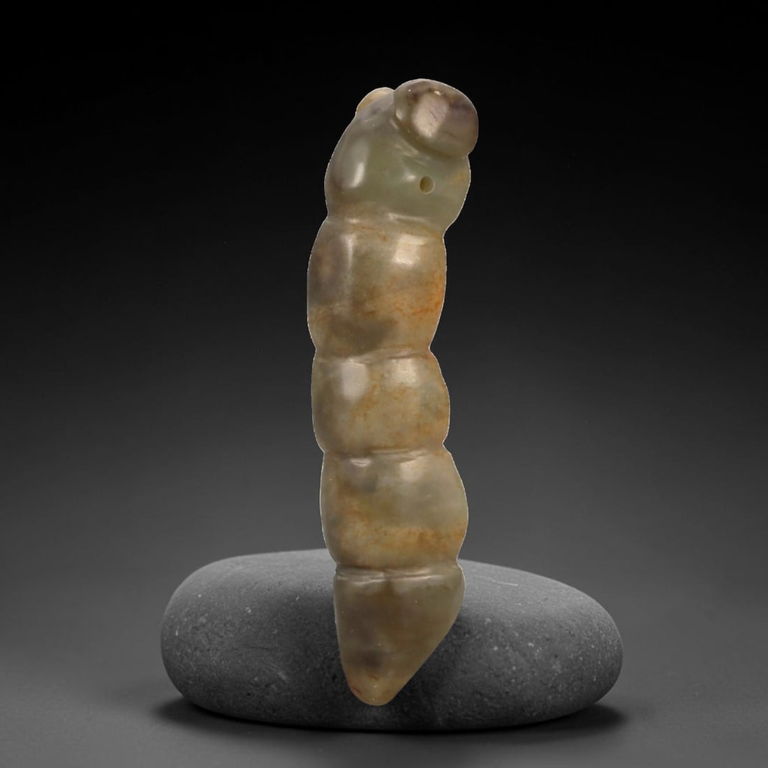 A Chinese Jade Worm Carving Pendant, Late Qing Dynasty, 64FB: Carved from mottled jade with grey-green tones and areas of brownish mineral inclusions, the object is modeled in the round as a stylized worm or larval form. The body is elongated and vertically orie