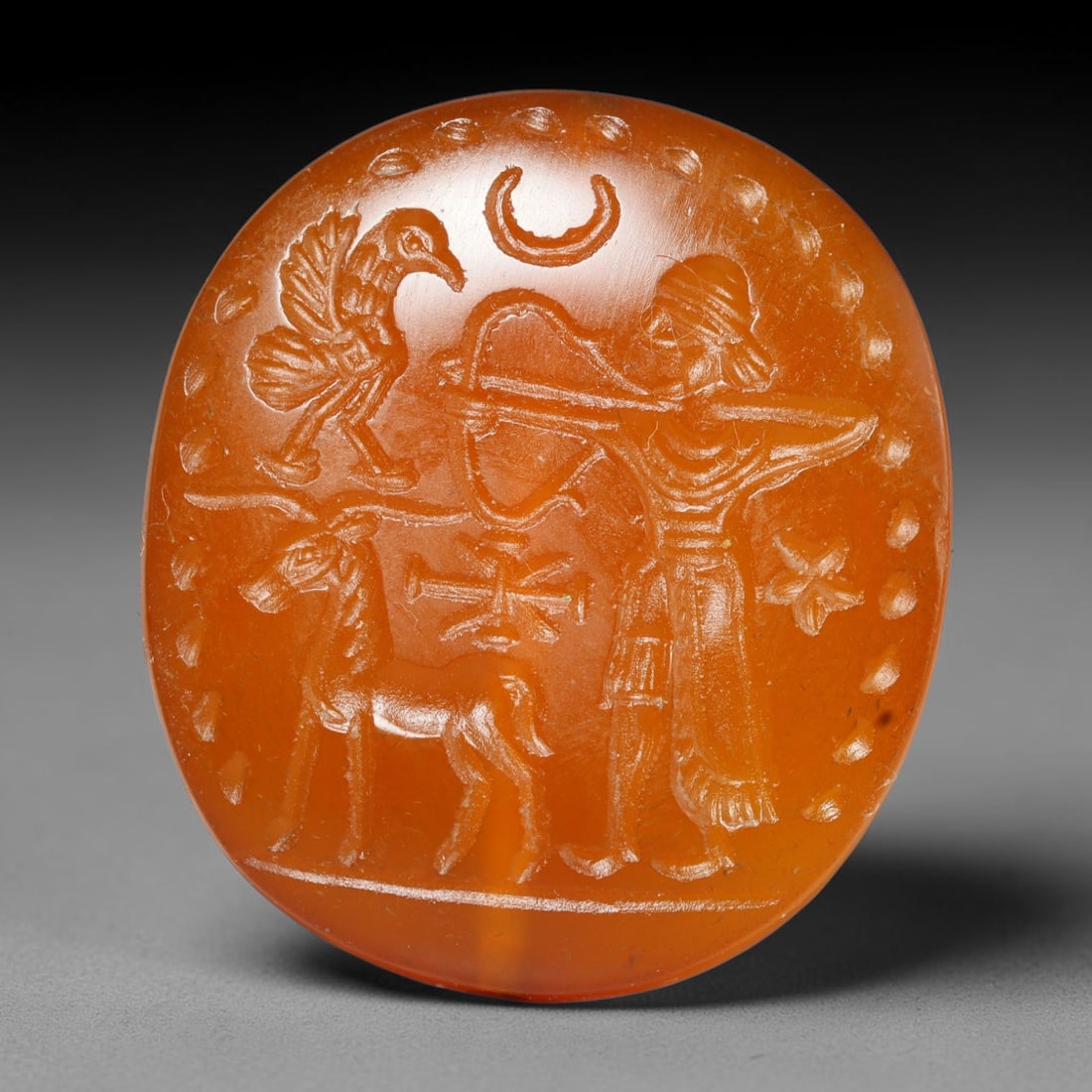 Ancient Medieval Early Islamic Period Carnelian Intaglio Bead (c.7th-11th Century CE), 79EC: This oval pierced bead is crafted from natural carnelian, featuring a detailed intaglio scene depicting an archer alongside a stag, avian figure, crescent moon and symbolic emblems, a classic composit