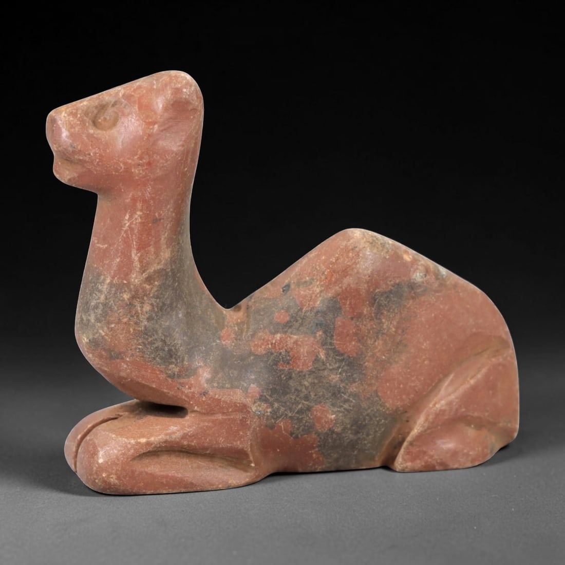Ancient Stone Camel Effigy - Near Eastern Animal Figurine, 47AM: This camel effigy is carved from solid reddish-brown stone with natural grey veining and mineral deposits, depicting a stylized camel in a recumbent, crouched posture. The piece features clean, incise
