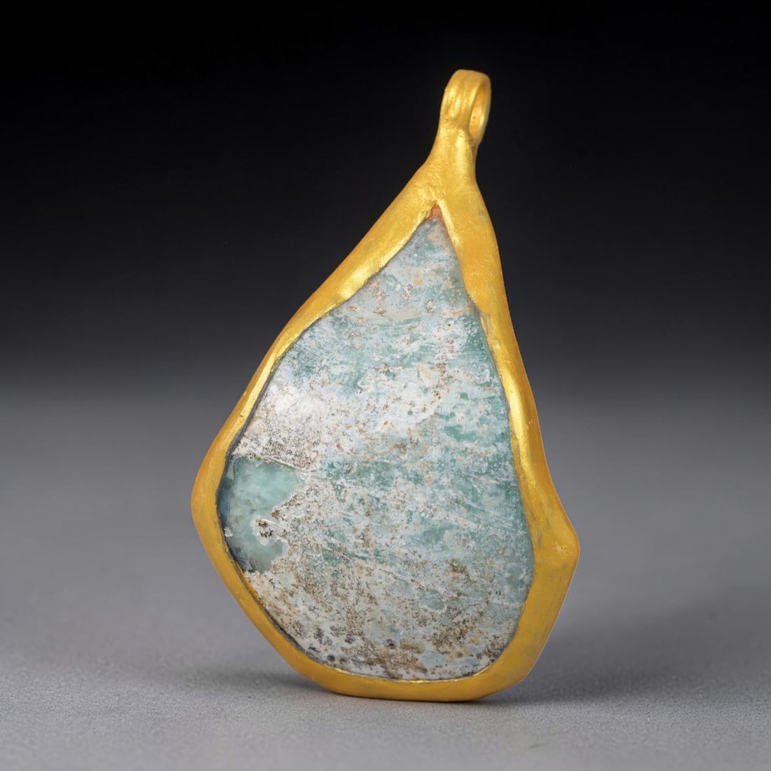 Ancient Roman Glass Slag in Modern Gilt-Edge Pendant, 28EH: This contemporary pendant is crafted from ancient Roman glass slag, featuring a fragmented, iridescent glass surface with weathered patina. The irregular form is bordered by a subtle gilt frame, added