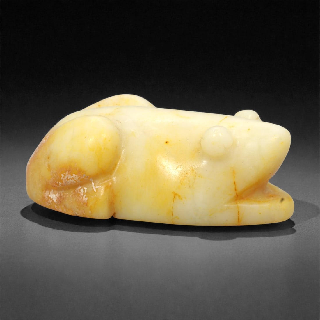 A Chinese Hetian Nephrite Jade Frog Figure, 47EC: Carved from pale creamy-white Hetian jade with areas of natural yellow and russet staining, the figure is modeled as a recumbent frog shown resting with its body flattened close to the ground. The hea