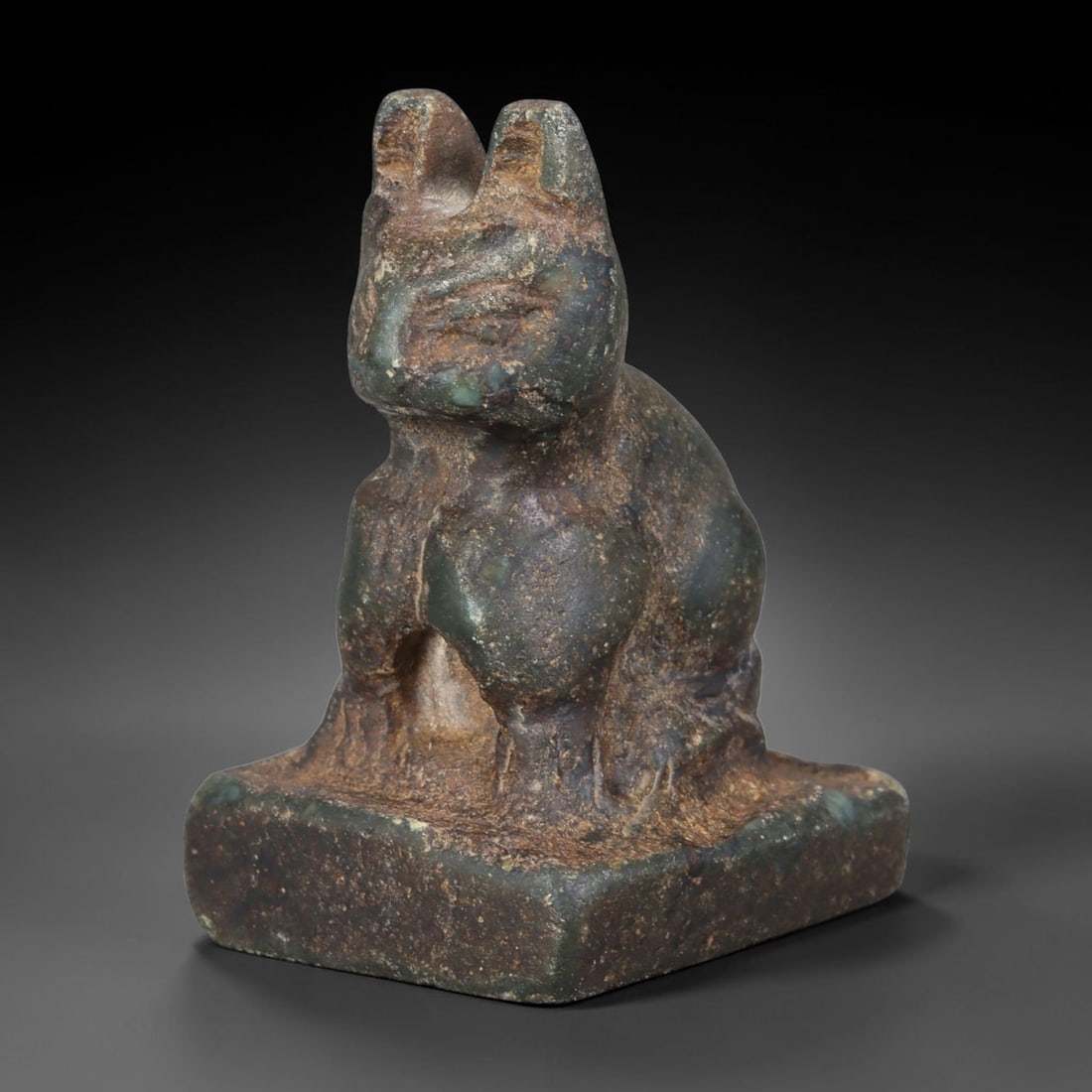 Ancient Egyptian Dark Green Stone Bastet Cat Amulet (664-30 BCE), 39AB: Hand-carved mottled dark green stone seated cat figurine representative of Bastet, mounted to a solid integrated rectangular plinth base. Subtle incised lines define facial features, ears and chest fu