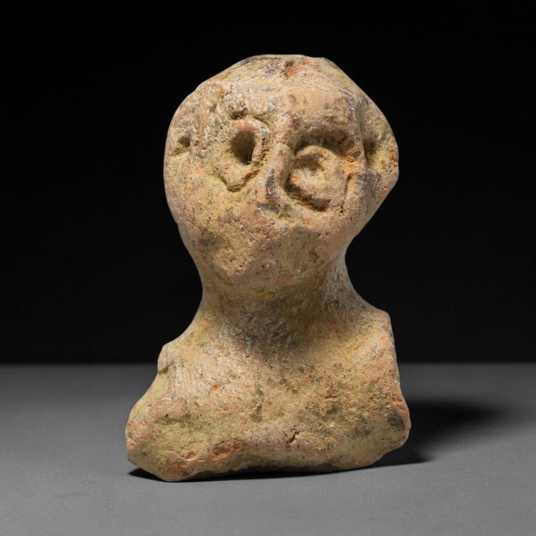 Ancient Hellenistic Terracotta Human Bust Fragment, 66ED: This weathered terracotta human bust fragment originates from the Hellenistic period. The simplified facial features are modelled with pierced circular eyes and minimal facial detailing, characteristi