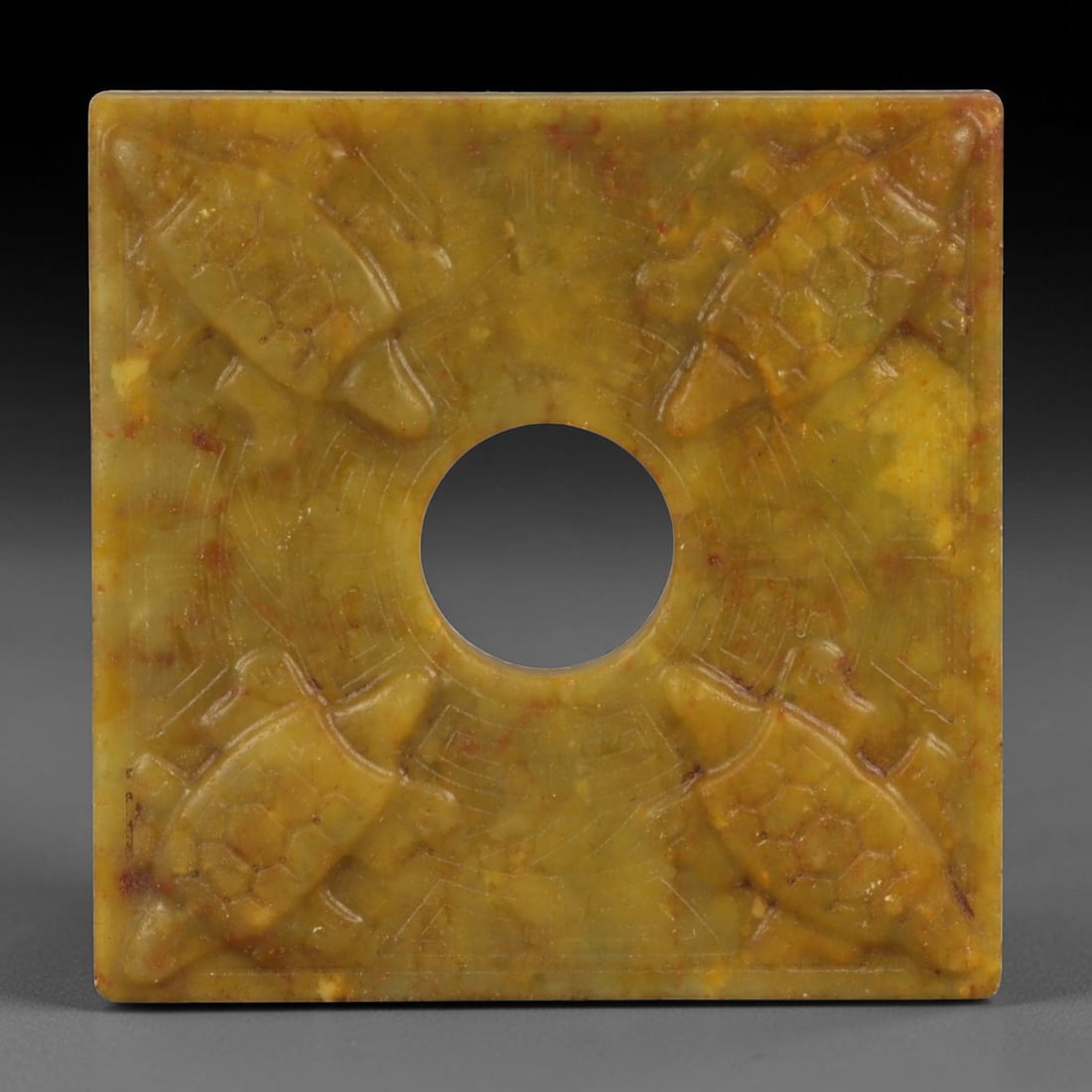 A Chinese Hardstone Plaque with Central Aperture, 77FD: A square hardstone plaque carved from mottled yellow-brown stone with russet inclusions and subtle surface translucency. The plaque is centered by a circular aperture, around which the surface is work