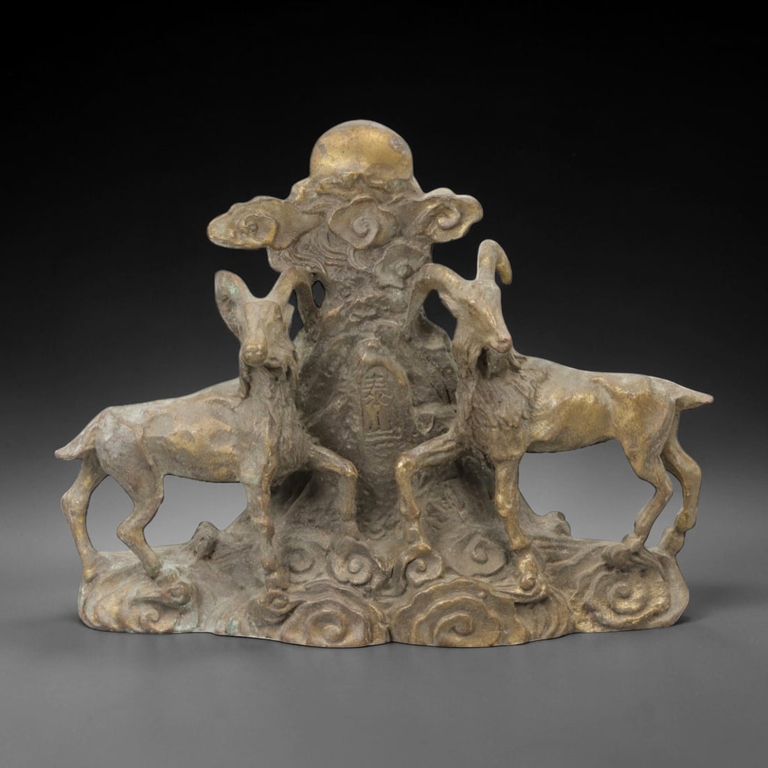 A Chinese Bronze Ram Group, 20th Century, 20FH: Cast in bronze with a dark brown surface and areas of lighter wear, the sculpture depicts a symmetrical group of two rams arranged on either side of a central vertical element. Each ram is shown stand