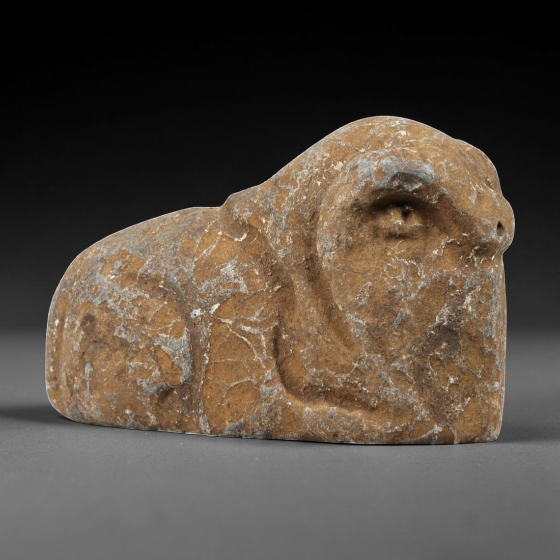 Ancient Stone Ram Effigy - Near Eastern Zoomorphic Figurine, 97FX: Carved from a weathered, veined stone, this effigy depicts a recumbent ram in a compact, stylized form. Relief details define the curled horns, snout, and rounded body, emphasizing the animal's robust