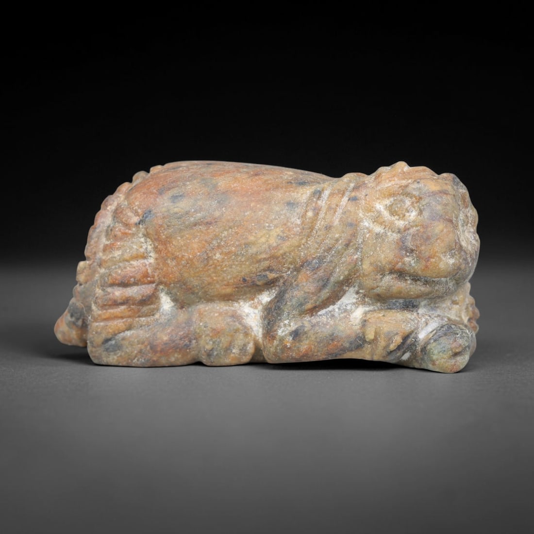 Ancient Sasanian Persian Recumbent Lion Effigy Amulet (224-651 CE), 5ER: This miniature votive talisman is carved from mottled hardstone originating from the Sasanian Persian Empire, shaped as a recumbent resting lion with stylized facial detailing, incised mane and simpli