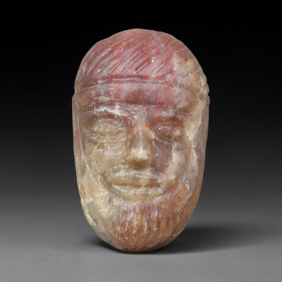 Ancient Sasanian Persian Banded Agate Human Head Amulet (224-651 CE), 8ER: This carved wearable amulet is fashioned from translucent banded agate originating within the Sasanian Persian Empire. Sculpted in the round as a full frontal male bearded human head, the piece displa
