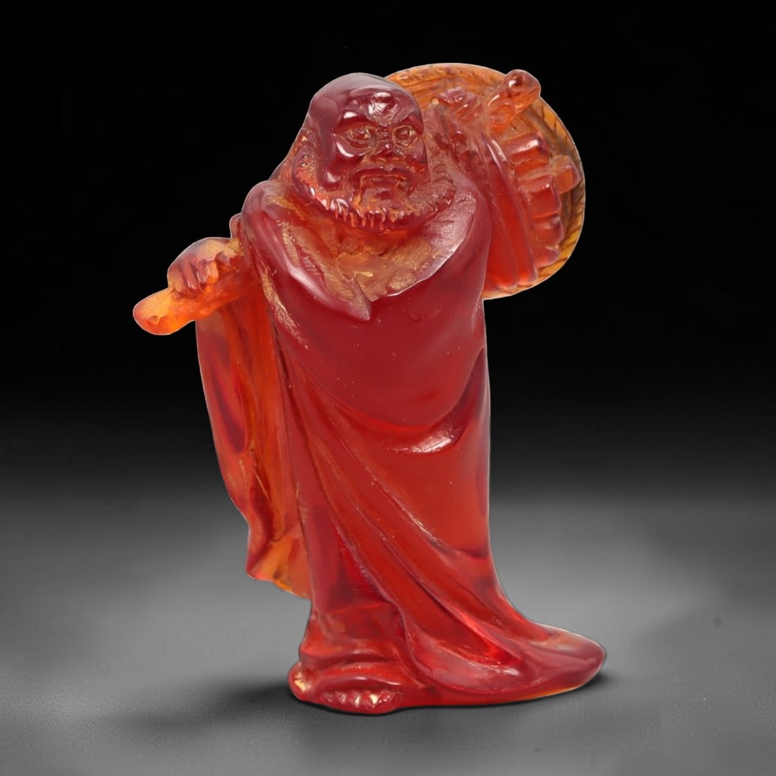 A Chinese Amber-Resin Figure of a Luohan, 41FH: Cast or carved from translucent amber-colored red resin with internal tonal variation, scattered inclusions, and minor entrapped bubbles, the figure represents a standing luohan shown holding a staff 