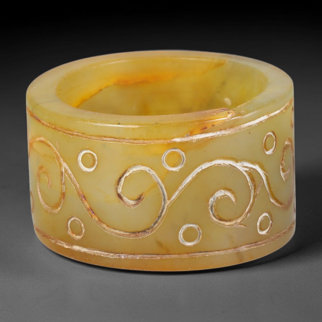A Chinese Nephrite Jade Cylinder Scroll Incised Decorative Form, 10AE: Fashioned from warm honey-toned nephrite jade carrying natural russet veining and internal tonal variation throughout the stone matrix, this hollow cylindrical stone form features hand incised continu