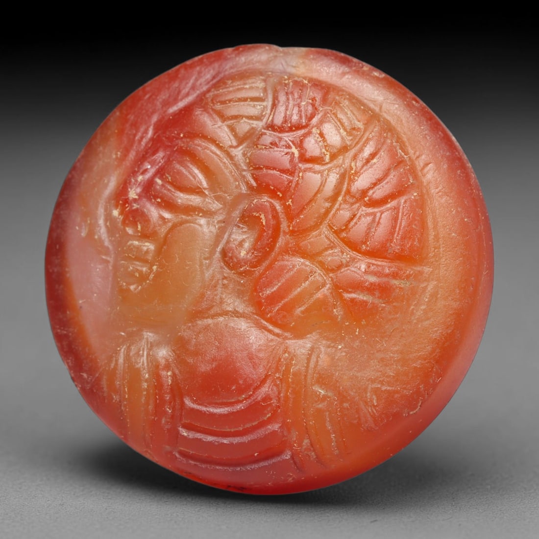 Ancient Medieval Early Islamic Period Carnelian Cameo Bead (c.7th-11th Century CE), 80EC: This circular pierced bead is crafted from natural carnelian, featuring a finely rendered cameo relief profile of an anthropomorphic bust with elaborate styled hair, a common motif within medieval Nea