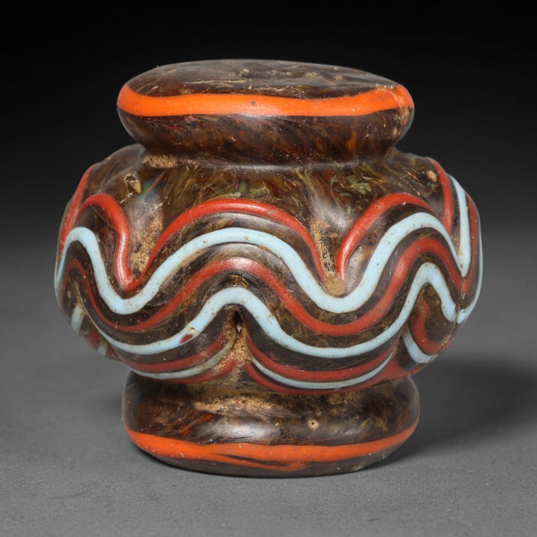 Ancient Medieval Islamic Wavy Trail Decorated Glass Bead (800-1200 CE), 72ED: This antique formed glass bead originates from the medieval Islamic glassmaking tradition, crafted for personal adornment and talismanic use. The marbled dark glass body features raised undulating tra