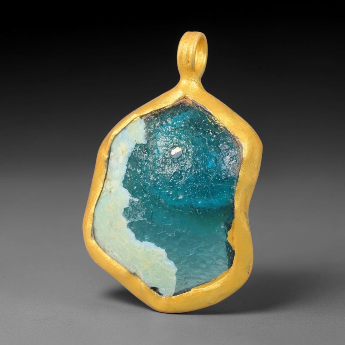 Ancient Roman Glass Slag in Modern Gilt-Edge Pendant, 30EH: This contemporary pendant is crafted from ancient Roman glass slag, featuring a fragmented, iridescent glass surface with weathered patina. The irregular form is bordered by a subtle gilt frame, added