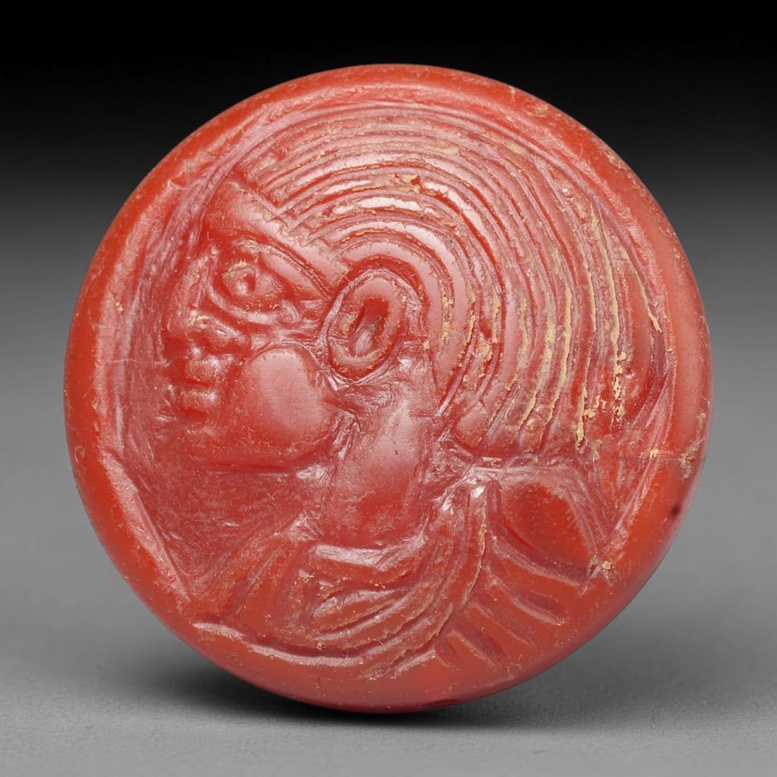 Ancient Medieval Early Islamic Period Carnelian Cameo Bead (c.7th-11th Century CE), 69EC: This circular pierced bead is crafted from natural carnelian, featuring a finely rendered cameo relief profile of an anthropomorphic bust with elaborate styled hair, a common motif within medieval Nea