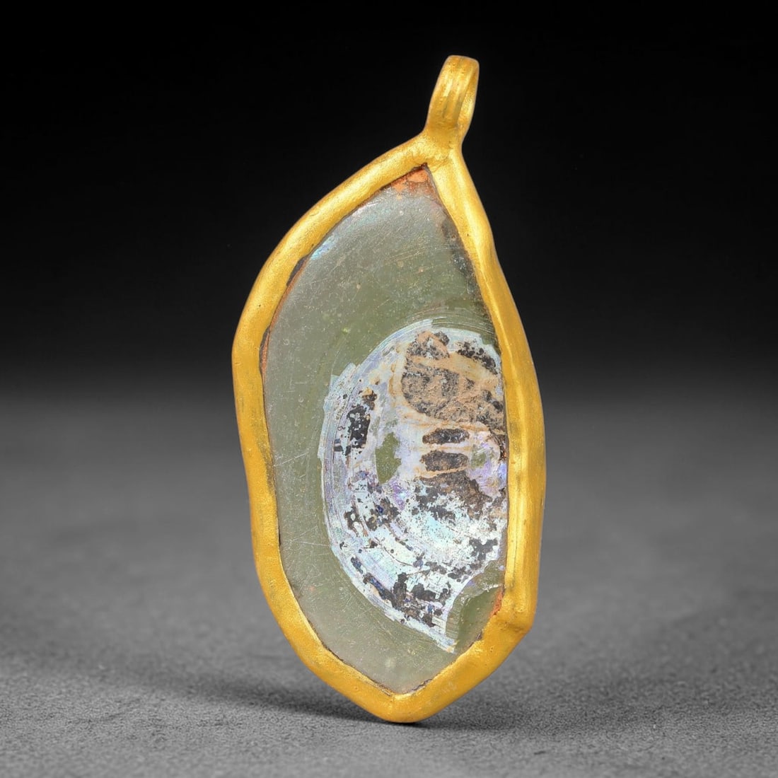 Ancient Roman Glass Slag in Modern Gilt-Edge Pendant, 70FY: This contemporary pendant is crafted from ancient Roman glass slag, featuring a fragmented, iridescent glass surface with weathered patina. The irregular form is bordered by a subtle gilt frame, added