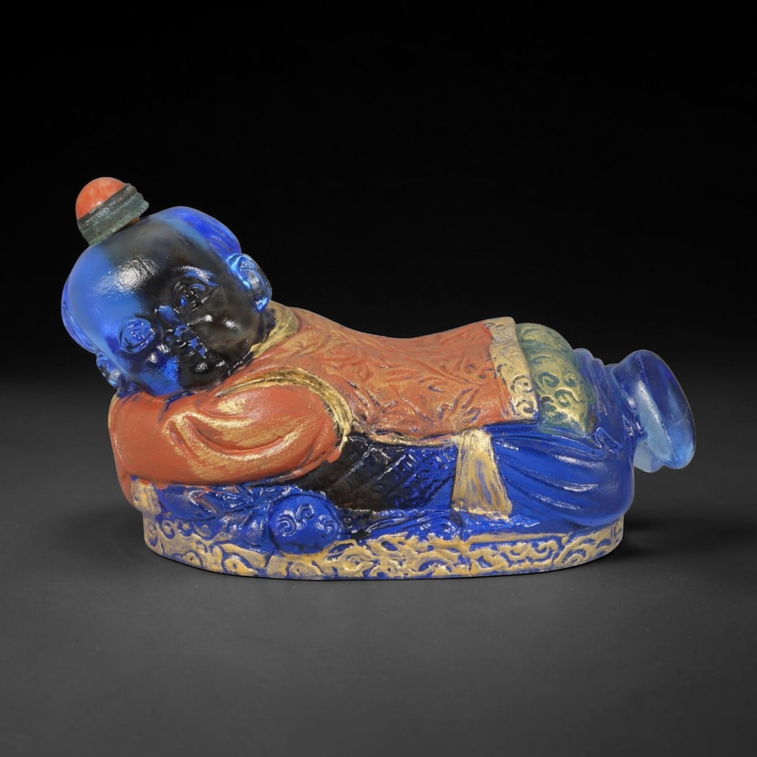 Vintage Chinese Liuli Glass Snuff Bottle - Reclining Sleeping Child, 46AS: This snuff bottle is moulded from translucent deep blue liuli glass, modelled as a reclining sleeping child figure. The robe features raised relief patterns finished with gilded and multi-coloured ena
