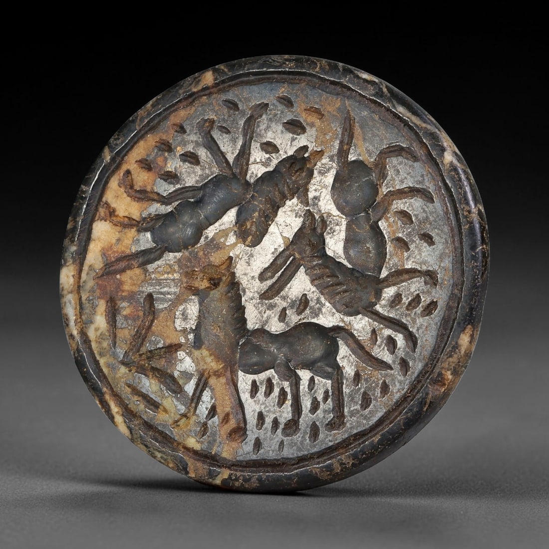 Ancient Syro-Hittite Dark Stone Intaglio Plaque (1200-800 BCE), 21EF: This circular stone intaglio plaque is carved from mottled dark stone, originating from Iron Age Syro-Hittite civilizations of Northern Syria and Eastern Anatolia. The surface is engraved with a compo