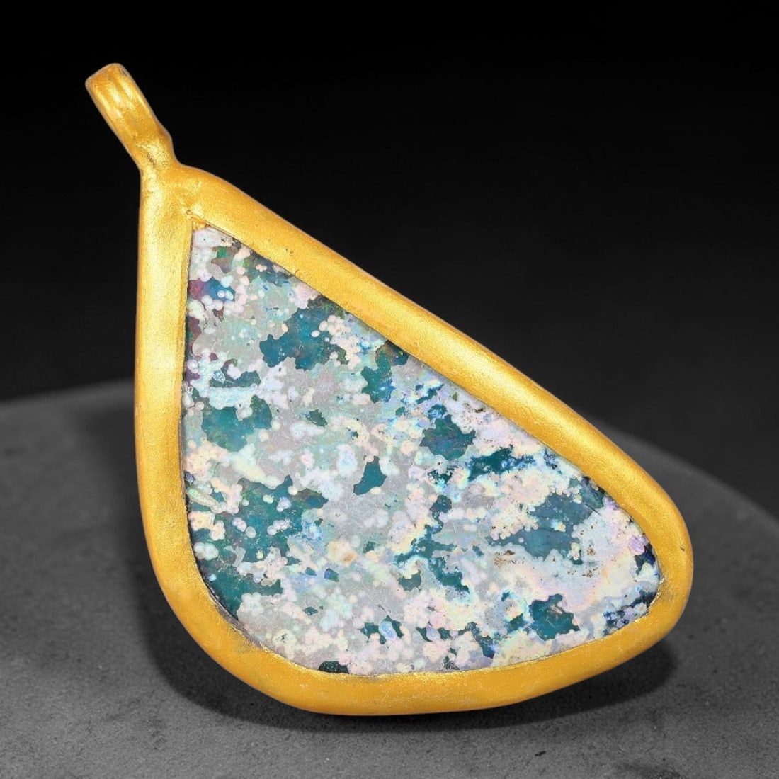 Ancient Roman Glass Slag in Modern Gilt-Edge Pendant, 62FY: This contemporary pendant is crafted from ancient Roman glass slag, featuring a fragmented, iridescent glass surface with weathered patina. The irregular form is bordered by a subtle gilt frame, added