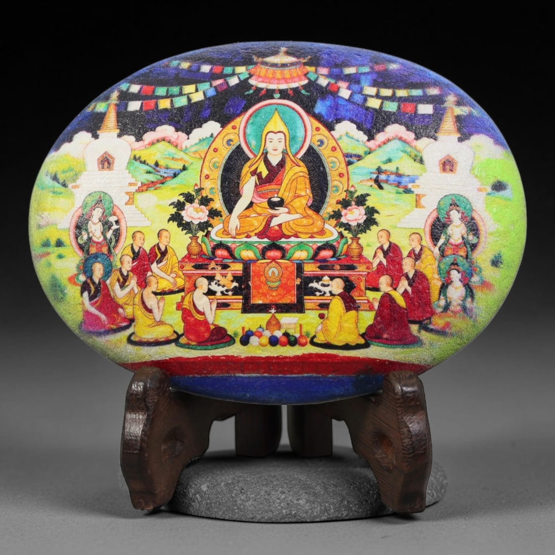 A Tibetan Thangka Style Painted River Stone with a Monastic Figure, 74BC: Painted on a naturally rounded river stone, the work depicts a central monastic figure in a thangka-style composition. The figure is shown seated in a meditative posture upon a lotus throne, wearing r