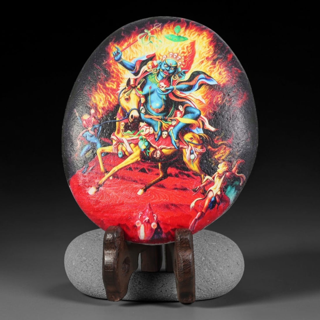 A Tibetan Thangka Style Painted River Stone with a Protective Deity, 64BC: Painted on a naturally rounded river stone, the work depicts a protective deity in a thangka-style composition. The figure is shown with a blue-grey countenance, riding a yellow horse and surrounded b