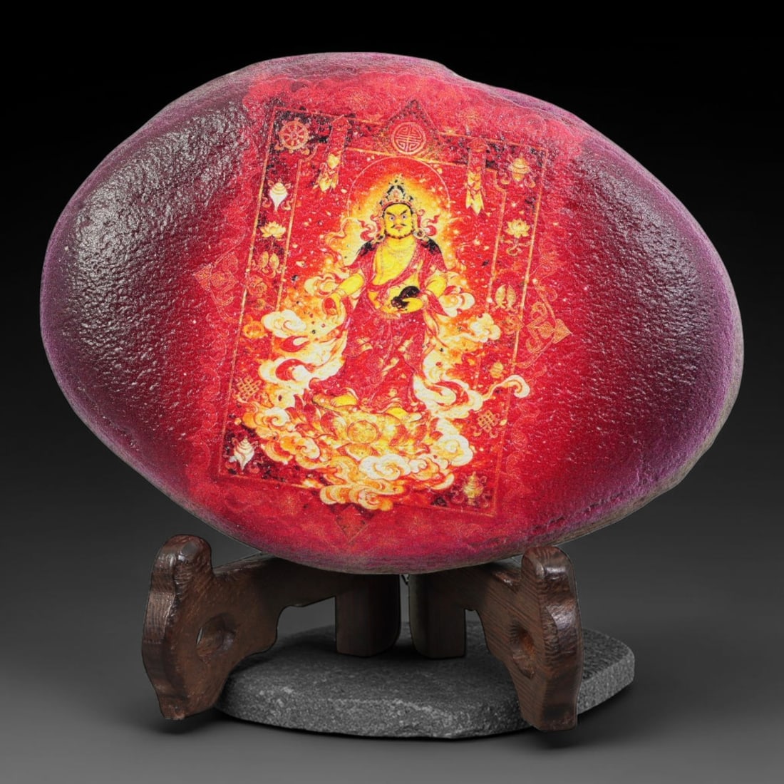 A Tibetan Thangka Style Painted River Stone with a Wealth Deity, 75BC: Painted on a naturally rounded river stone with red veining, the work depicts Yellow Jambhala, a wealth deity, in a thangka-style composition. The figure is shown in a robust form with a serene expres