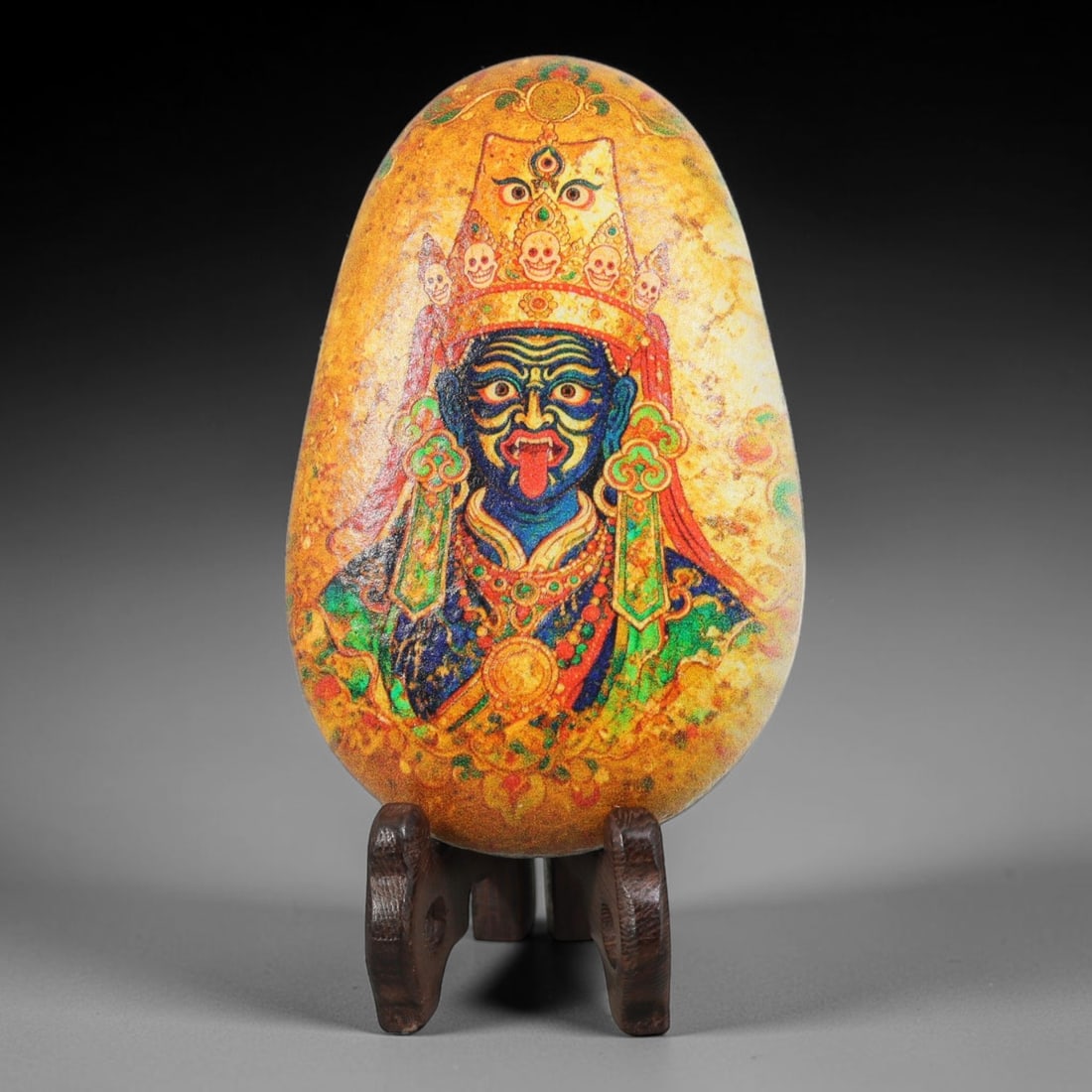 A Tibetan Thangka Style Painted River Stone with a Protective Deity, 81BC: Painted on a naturally rounded, teardrop-shaped river stone, the work depicts a protective deity in a thangka-style composition. The figure is shown with a blue-black countenance, wearing a crown deco