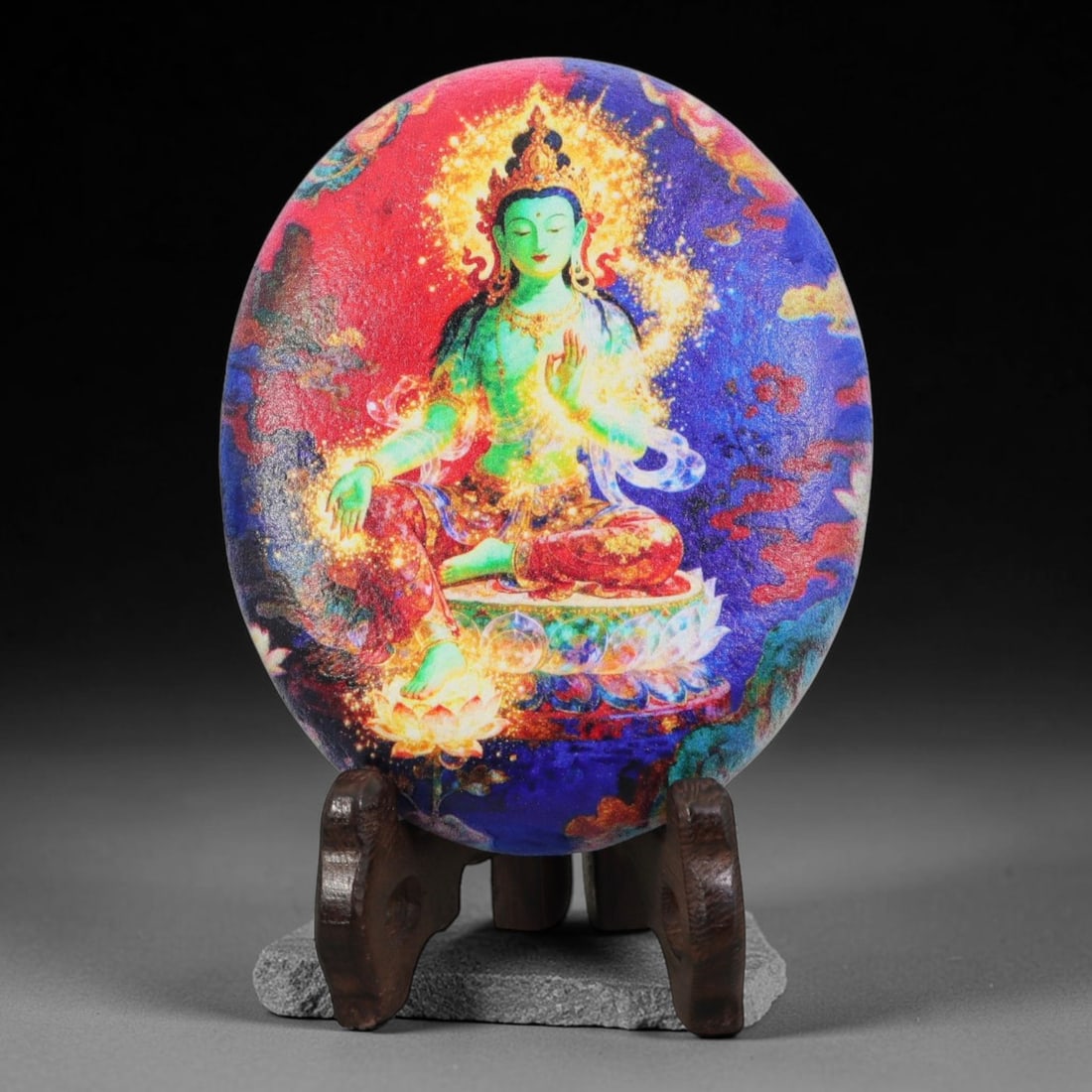 A Tibetan Thangka Style Painted River Stone with a Green Tara, 66BC: Painted on a naturally rounded river stone, the work depicts Green Tara, a female Buddhist deity, in a thangka-style composition. The figure is shown seated in a meditative posture upon a lotus base, 