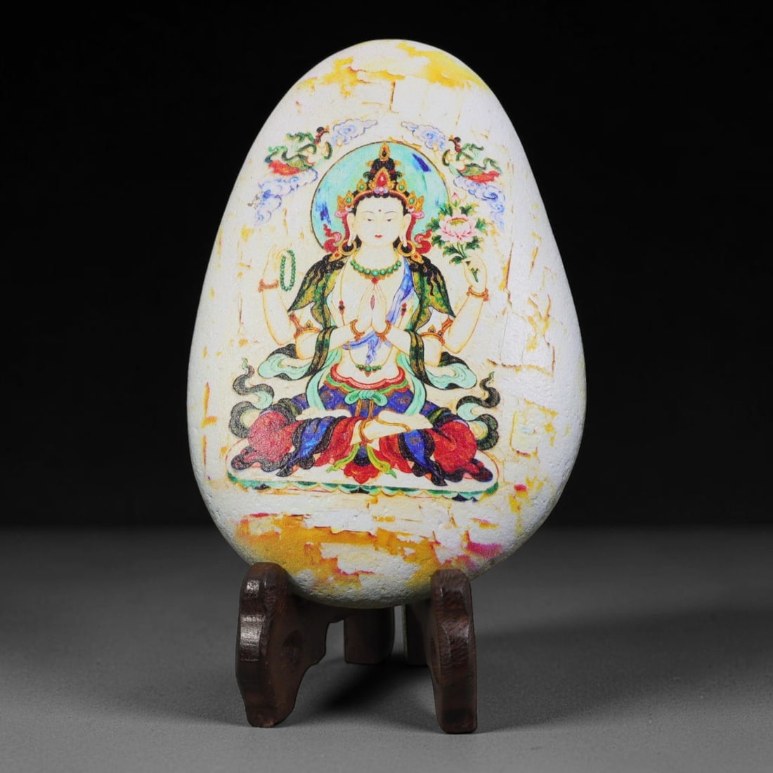 A Tibetan Thangka Style Painted River Stone with a Four-Armed Avalokiteshvara, 65BC: Painted on a naturally rounded, teardrop-shaped white river stone, the work depicts a four-armed Avalokiteshvara in a thangka-style composition. The figure is shown in a meditative posture, holding a 