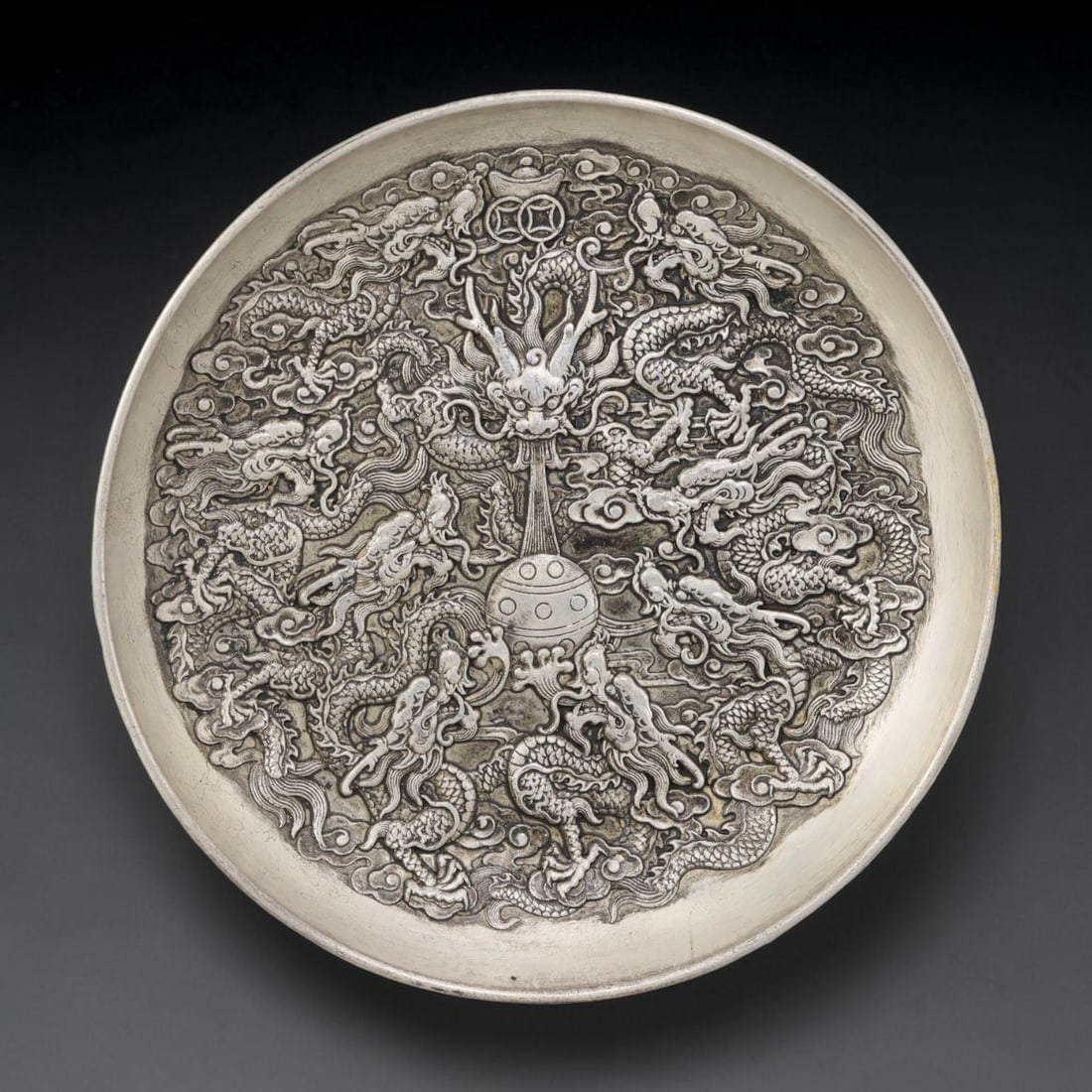 A Nine Dragons German Silver Decorative Plate, 29AD: A circular plate in German silver (a nickel-silver alloy), the center decorated in high relief with nine dragon motifs. The central dragon is shown with a fierce head, spouting a stream of water towar
