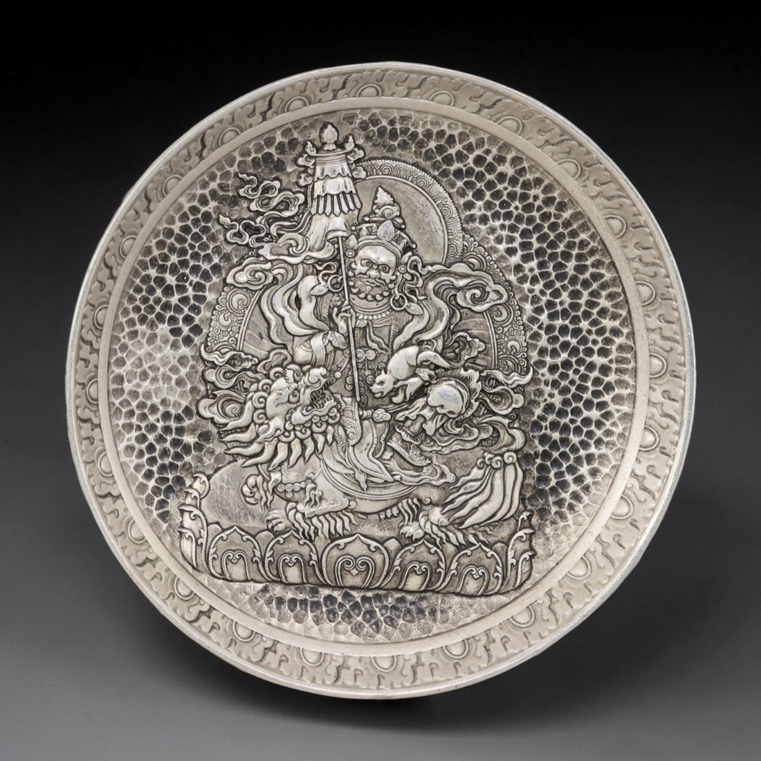 A Tibetan Wealth Deity German Silver Decorative Plate, 32AD: A circular plate in German silver (a nickel-silver alloy), the center decorated in high relief with a wealth deity, likely Vaishravana, in a Tibetan Buddhist manner. The deity is shown with a dignifie